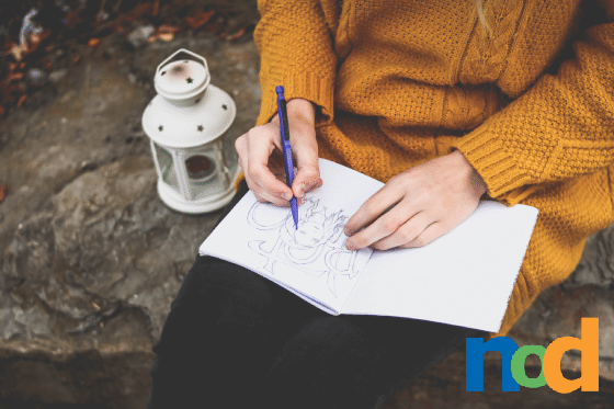 Woman sitting on a rock wearing oversized yellow sweater drawing in a sketchbook. There is a white lamp next to her with stars on it.