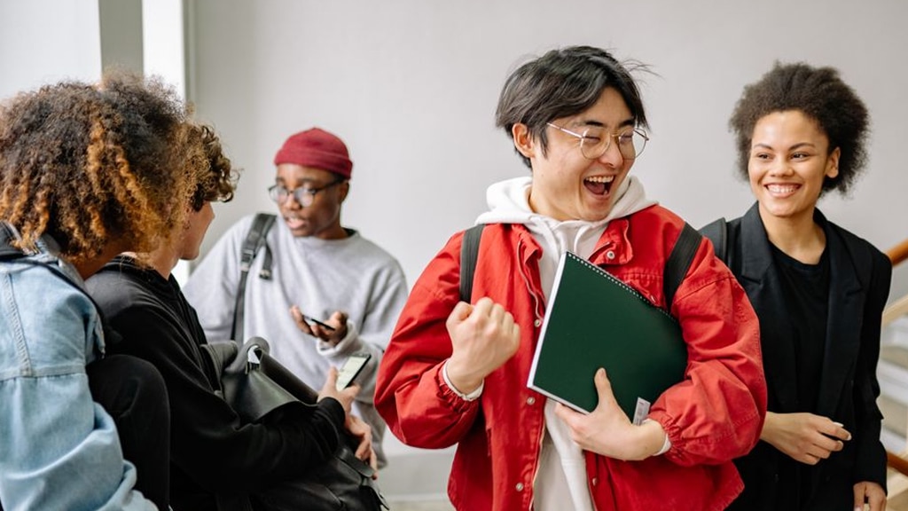 A diverse group of students chatting and smiling in a hallway, one person in a red jacket holding notebooks and folders.