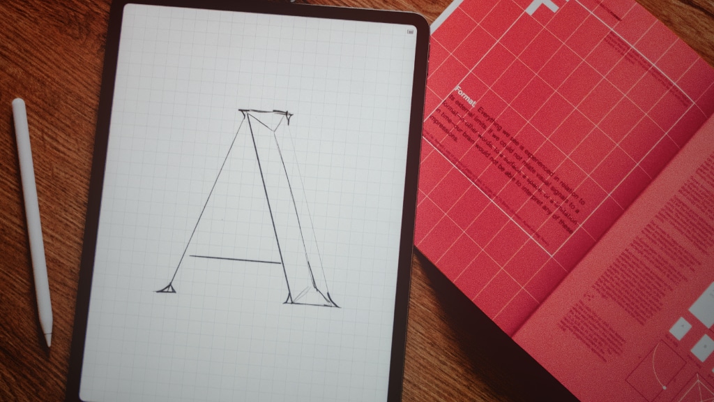 Tablet displaying a hand-drawn uppercase A on graph-paper background, with a stylus to the left and a red notebook on the right.