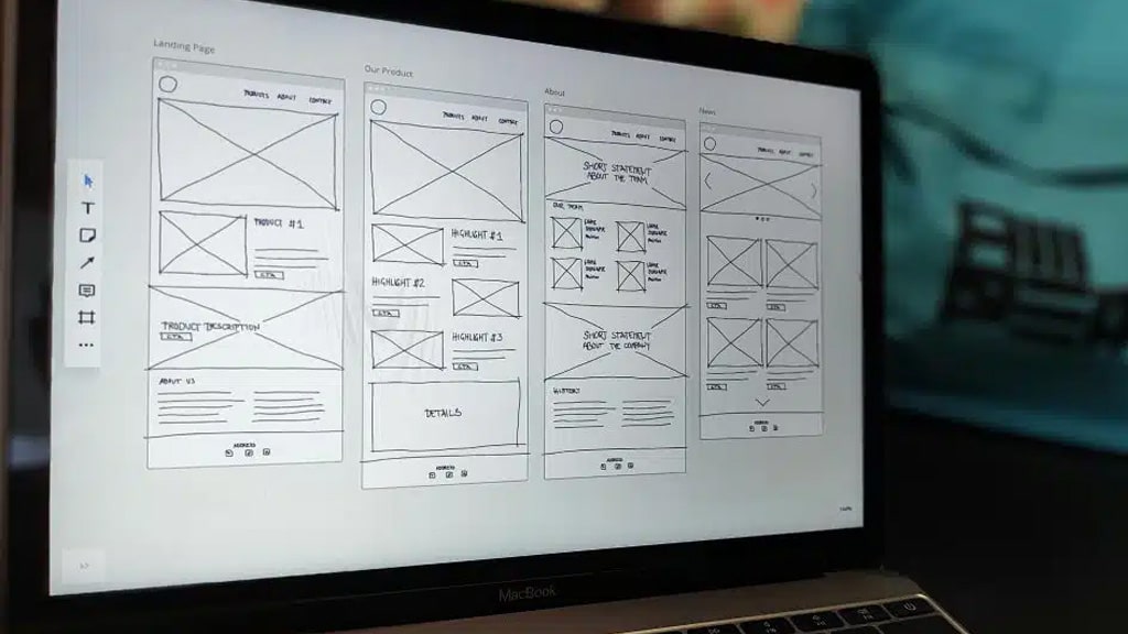 Laptop screen shows four-column website wireframe sketches with headers and placeholders on a design sheet on screen.